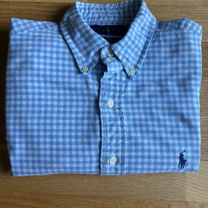 Blårutig skjorta från Ralph Lauren - Säljer en blårutig skjorta från Ralph Lauren i slim fit-modell. Skjortan är tillverkad i 100% bomull och har långa ärmar, klassisk krage och knappar framtill. Ikoniska Polo-loggan broderad på bröstet. Perfekt för en stilren look. Tyvärr så finns det små svett fläckar som knappas syns men de finns.