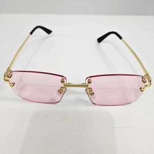 Rosa rektangulära solglasögon med gulddetaljer - Trendiga solglasögon med rektangulära rosa glas och tunna guldfärgade bågar. Glasögonen har en unik gulddetalj vid bryggan och svarta skalmtippar. Perfekt accessoar för att sticka ut med en färgklick.