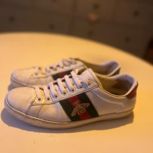 2st par Vita sneakers med broderad bi och röd/grön rand - Säljer ett par vita sneakers från GG med den klassiska röd-gröna randen på sidan och en broderad guldfärgad bi. Skorna har rund tå, platt sula och detaljer i rött och grönt på hälen.