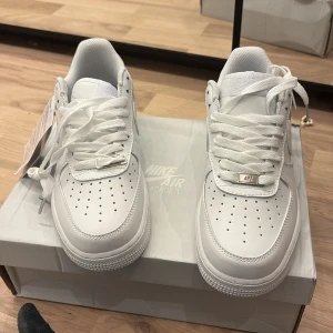 Nike Air Force 1 vita sneakers - Klassiska helvita Nike Air Force 1 sneakers med perforerad tå, platt sula och snörning. Skorna har en stilren design med rund tå och är tillverkade i syntetmaterial. Perfekta för dig som gillar en clean och tidlös look.