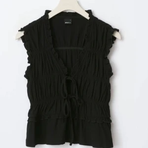 frill tie front top - Svart ärmlös topp från Gina Tricot med volangkant och knyt framtill. Toppen har en v-ringad design och smockade detaljer som ger en feminin och trendig look. jätte fin topp men passade inte mig i storleken. Ny pris 280 kr. pris kan diskuteras. säljer en lika dan i vit på mitt konto❤️
