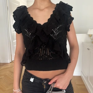 Svart volangtopp från Zara - Säljer en svart topp från Zara med volangdetaljer och virkade partier. Toppen har korta ärmar och en djup v-ringning både fram och bak. Perfekt för dig som gillar romantiska detaljer och en feminin stil. Den är väldigt eftertraktad därav pris. Passar även s