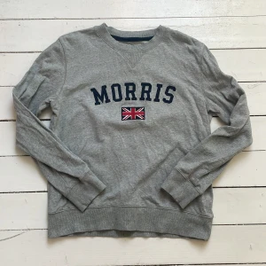 Grå sweatshirt från Morris - Säljer en klassisk grå sweatshirt från Morris med broderad logga och brittisk flagga på bröstet. Tröjan har rund halsringning, långa ärmar och ribbade muddar vid ärmslut och nederkant. Perfekt för en stilren och avslappnad look.