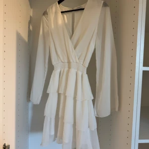 Perfekta studentklänningen  - Säljer en vit omlottklänning med långa ärmar och flera lager volanger i kjolen. Klänningen har v-ringning och markerad midja med resår. Perfekt för sommarens tillfällen! Den är från Bubbleroom