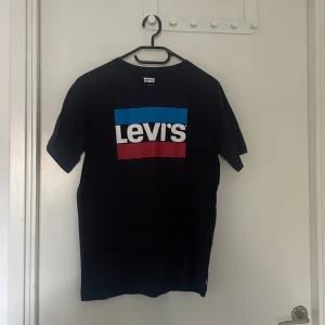 Svart t-shirt från Levi's med logga - Säljer en svart t-shirt från Levi's med den klassiska stora loggan i vitt, blått och rött framtill. T-shirten har rund hals och korta ärmar. Helt oanvänd och har storleken S. Priset kan diskuteras.