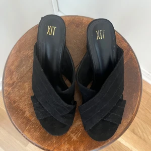 Svarta klackskor från XIT storlek 39 - Snygga svarta klackskor från XIT med korsade remmar i mocka. Skorna har en öppen tå och en stabil klack som ger en elegant look. Sparsamt använda!