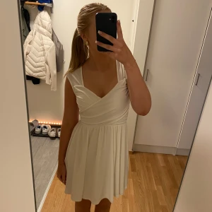 Vit omlottklänning - Säljer en söt vit omlottklänning med v-ringad design. Klänningen är ärmlös och har en kort längd, perfekt för sommarens varma dagar. Den har en elegant och enkel stil som passar till många tillfällen.