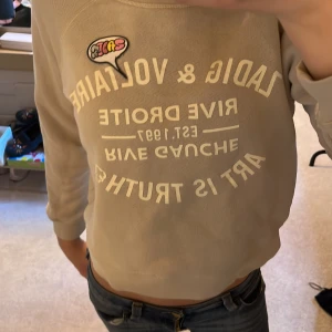 Beige tröja från Zadig & Voltaire - Säljer en beige tröja från Zadig & Voltaire med tryck framtill och en färgglad detalj. Tröjan har långa ärmar och rund halsringning. Perfekt för en avslappnad stil.