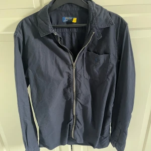 Polo Ralph Lauren skjorta - Säljer en stilren mörkblå skjorta från Polo Ralph Lauren med dragkedja framtill. Skjortan har en bröstficka och långa ärmar, perfekt för en avslappnad men snygg look inför våren😍