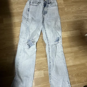 Ljusa jeans med slitningar - Säljer ett par ljusa jeans med coola slitningar på knäna och längs ner. De har en klassisk femficksdesign och knappgylf. Perfekta för en avslappnad stil.