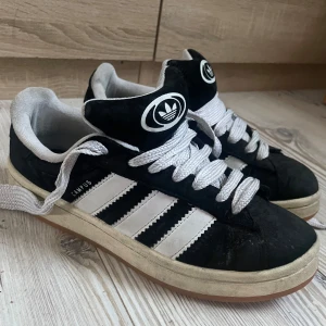 Svarta Adidas Campus sneakers - Säljer ett par klassiska svarta Adidas Campus sneakers med vita detaljer och snörning. Är lite smutsiga men tror verkligen att de går att tvätta upp!!