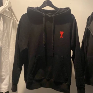 Svart hoodie från Ami - Säljer en stilren svart hoodie från Ami med en röd broderad logotyp på bröstet. Tröjan har en klassisk passform med långa ärmar och en praktisk magficka. Perfekt för en avslappnad look.