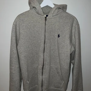 Grå hoodie från Polo Ralph Lauren - Säljer en stilren grå hoodie från Polo Ralph Lauren med dragkedja och det klassiska logotypbroderiet på bröstet. Tröjan har en bekväm passform och är perfekt för vardagsbruk. Den har en praktisk huva och fickor framtill. Ett litet märke på armen (se bild)