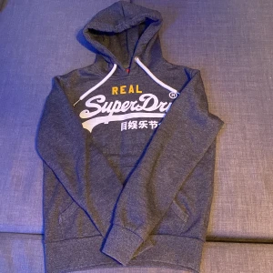 Superdry Hoodie - Säljer denna riktigt snygga mörkgråa Superdry hoodie! Har blivit använd fåtal gånger och den är i mycket bra skick. Inga defekter!