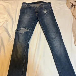 Blå jeans från Nudie Jeans 29x32 - Säljer ett par blå Nudie Jeans med slitna detaljer och en klassisk femficksdesign. Jeansen har en normal passform och är tillverkade i denim med en snygg tvätt. Perfekta för en avslappnad stil.