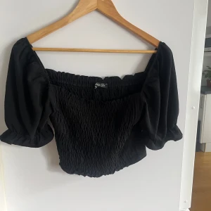 Svart offshoulder topp från Boohoo - Mycket snygg svart offshoulder topp från Boohoo. Toppen har puffiga ärmar och en smockad design som ger en fin passform. Perfekt för att styla med både kjol och jeans. Hör av dig vid intresse!🌺