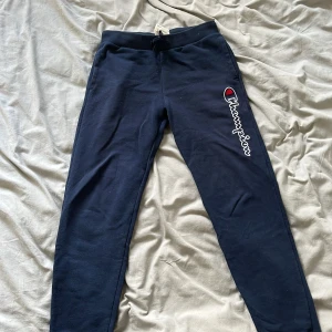Mörkblå mjukisbyxor från Champion - Säljer ett par mörkblå mjukisbyxor från Champion med logotypen tryckt längs benet. Byxorna har en snörning i midjan för justerbar passform och är perfekta för en avslappnad stil. 13-14 år