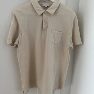 Beige pikétröja från Sunspel - Säljer en stilren beige pikétröja från Sunspel. Tröjan har korta ärmar och en klassisk krage med två knappar. Den är tillverkad i ett bekvämt material och har en bröstficka för extra stil. Perfekt för en avslappnad men ändå snygg look.