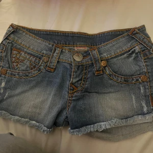 Jeansshorts från True Religion - Säljer ett par snygga jeansshorts från True Religion. De är blå med fransiga kanter och har coola detaljer som kontrastsömmar och en broderad bakficka. Perfekta för sommardagar! 🌞