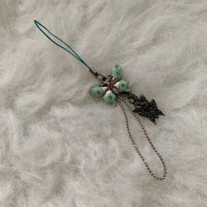 Fjärilsformad mobilaccessoar med kedja - Charmig mobilaccessoar med en fjärilsformad design i mintgrön och silver. Dekorerad med rosa och gröna stenar samt en liten kedja med extra fjärilsdetaljer. Perfekt för att ge din mobil en personlig touch.