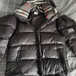 Svart dunväst från Burberry - Snygg svart dunväst från Burberry med avtagbara ärmar och dragkedja. Den har en quiltad design och en huva med klassiskt mönstrat foder. Perfekt för kyliga dagar när du vill hålla stilen.