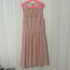 Beige rosa klänning med spetsdetaljer - Elegant beige rosa klänning med vacker spets upptill och en luftig kjol. Den är ärmlös och har en rund halsringning. Perfekt för sommarens festligheter.