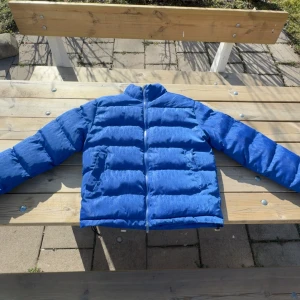 Blå pufferjacka från Dior - Säljer en snygg blå pufferjacka från Dior. Jackan har en quiltad design med långa ärmar och en dragkedja framtill. Perfekt för kyligare dagar och ger en stilren look. Den har ett diskret mönster och en logotyp på ärmarna, har använt jackan  ett fåtal gånger och är ett unik jacka som är perfekt för en person som har bra stil😀