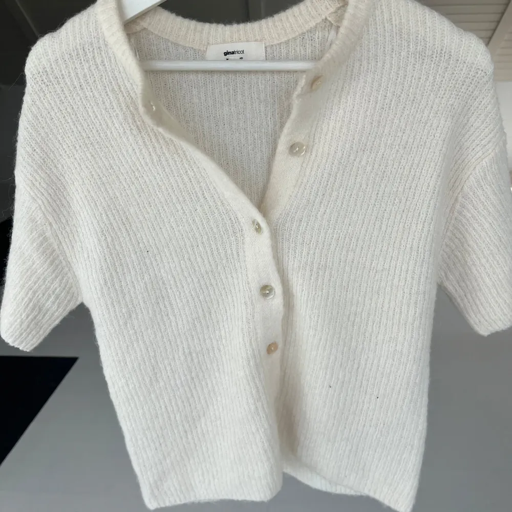 Säljer en stilren vit stickad kofta från Gina Tricot. Koftan har korta ärmar och knappar framtill, perfekt för en avslappnad look. Den är mjuk och bekväm, idealisk för både vår och sommar. Passar perfekt att styla med ett vitt linne under! Nyskick! . Neuletakit & Villapaidat.