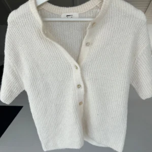 Vit stickad kofta från Gina Tricot - Säljer en stilren vit stickad kofta från Gina Tricot. Koftan har korta ärmar och knappar framtill, perfekt för en avslappnad look. Den är mjuk och bekväm, idealisk för både vår och sommar. Passar perfekt att styla med ett vitt linne under! Nyskick! 