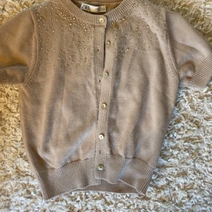 Beige kofta från Zara - Säljer en söt beige kofta från Zara med korta ärmar och pärldetaljer vid halsringningen. Koftan har knappar framtill och ribbade kanter. Perfekt för en stilren look.