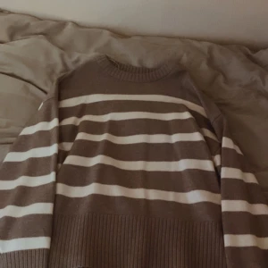 Grå och vit randig tröja - Säljer en mysig grå och vit randig tröja med ribbade muddar och nederkant. Perfekt för kyliga dagar. Tröjan har långa ärmar och en rund halsringning och slits.