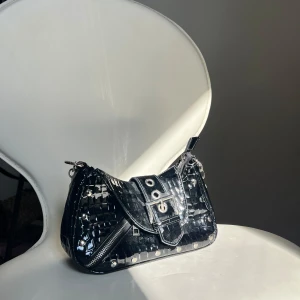 Svart axelväska med kedjedetaljer från NAKD - Snygg svart axelväska med glansig finish och krokodilmönster. Väskan har en justerbar axelrem med kedjedetaljer och en dekorativ bältesdetalj på framsidan. Perfekt för att ge din outfit en edgy touch. 