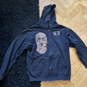 Svart hoodie med tryck - Säljer en svart hoodie med ett unikt tryck av en maskerad figur på framsidan och text på baksidan. Hoodien har dragkedja och en klassisk känguruficka framtill. Perfekt för en cool och avslappnad stil.