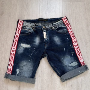 Jeansshorts från Mario Morato - Snygga jeansshorts från Mario Morato med slitna detaljer och uppvikta ben. De har en cool röd och vit logotypremsa längs sidorna och klassiska femficksdesign. Perfekta för en avslappnad stil. helt nya är ej använda.😀