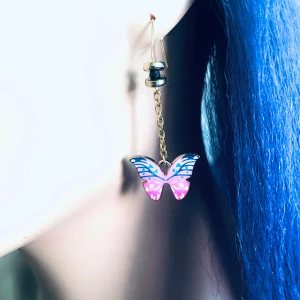 By prisförslag- 1 st Fjärilsörhänge i rosa och blått - Charmigt örhänge med hängande fjärilar i rosa och blått. Den har en tunn kedja och en liten svart pärla nära örat. Perfekta för att lägga till en färgglad touch till din stil.