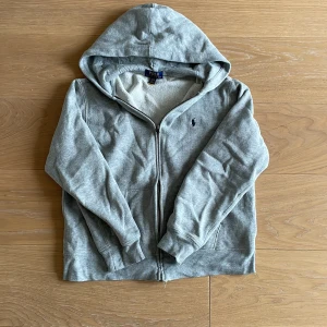Grå ziphoodie från Polo Ralph Lauren - Säljer en stilren grå hoodie från Polo Ralph Lauren med dragkedja och broderad logga på bröstet eftersom den blivit för liten för mig. Tröjan har en mysig huva och praktiska fickor framtill. Perfekt för en avslappnad look! Skriv för frågor eller mer bilder 💯💯