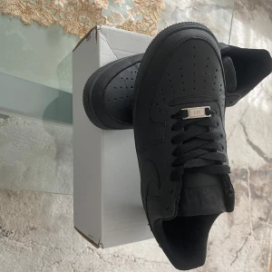 Svarta Nike Air Force 1 - Säljer ett par klassiska svarta Nike Air Force 1 sneakers. Skorna har en stilren design med perforerade detaljer på tån och en robust sula. Perfekta för en casual look.
