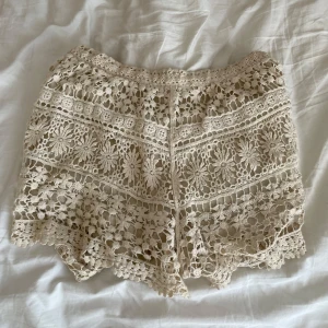 Beige spets shorts - Säljer ett par snygga beige shorts med vacker spetsdesign. Perfekta för en somrig look med sitt luftiga och stilrena mönster. De har en elastisk midja för extra komfort. Passar perfekt till en avslappnad dag i solen.