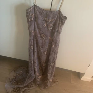 Långklänning med broderade detaljer - Elegant långklänning i en dämpad lila nyans grå med vackra broderade blommor och paljettdetaljer. Klänningen har smala axelband och en flytande siluett som ger en romantisk känsla. Perfekt för speciella tillfällen. Tvättas med insidan ut