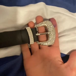 Svart bälte med glittrigt spänne från BikBok - Snyggt svart bälte från BikBok med ett glittrigt spänne som ger en extra touch av glamour. Perfekt för att lyfta vilken outfit som helst. Bältet är i storlek XS-S och har en klassisk design med flera hål för justering.