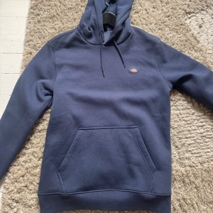 Mörkblå hoodie från Dickies - Säljer en stilren mörkblå hoodie från Dickies med en klassisk känguruficka och justerbar huva. Perfekt för en avslappnad stil. Den har långa ärmar och en liten logga på bröstet.
