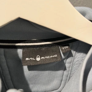 Grå jacka från Sail Racing - Säljer en stilren grå jacka från Sail Racing med dragkedja och broderad logga på bröstet. Jackan har en hög krage och en praktisk bröstficka med dragkedja. Äkta och ny 