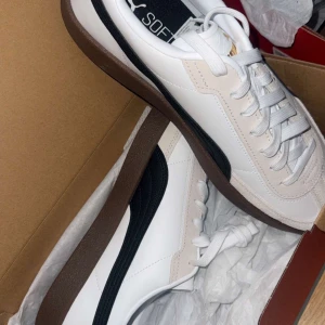 Vita och svarta sneakers från Puma - Snygga vita sneakers från Puma med svarta detaljer och brun sula. Skorna har snörning och en klassisk design som passar perfekt för vardagsbruk.