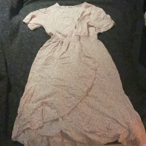 Ljusrosa blommig klänning - Ljusrosa klänning med blommigt mönster och omlott-design. Klänningen har korta ärmar och en v-ringad hals. Säljer då den är förstor Perfekt för vår och sommar!