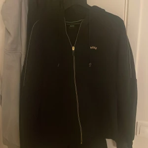 Svart hoodie från Boss - Säljer en stilren svart hoodie från Boss med dragkedja och huva. Den har diskreta vita detaljer längs ärmarna och en liten logotyp på bröstet. Perfekt för en avslappnad look.