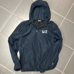 Mörkblå vindjacka från EA7 - Snygg mörkblå vindjacka från EA7 med huva och dragkedja framtill. Jackan har en stilren design med logotyp på bröstet och praktiska fickor med dragkedja. Perfekt för blåsiga dagar.