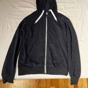 Mörkgrå hoodie från Divided - Säljer en stilren blå hoodie från Divided med dragkedja och vita snören i huvan.