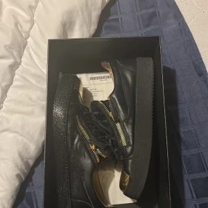 Giuseppe Zanotti - Snygga svarta sneakers med gulddetaljer och dragkedjor på sidorna. Skorna har en unik design med en guldfärgad tå och snörning framtill. Perfekta för att ge en stilren touch till din outfit.