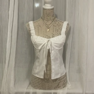 Vit korsettopp med knytband - Snygg vit korsettopp med breda axelband och knytband framtill. Passar utmärkt till sommaren. Är stretchig på baksidan så passar storlek 34-38. 