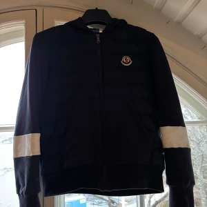 Svart jacka med huva och dragkedja - Snygg svart jacka från Moncler med huva och dragkedja. Jackan har vita detaljer på ärmarna och ett broderat märke på bröstet. Perfekt för kyligare dagar. Den är storlek 14 år